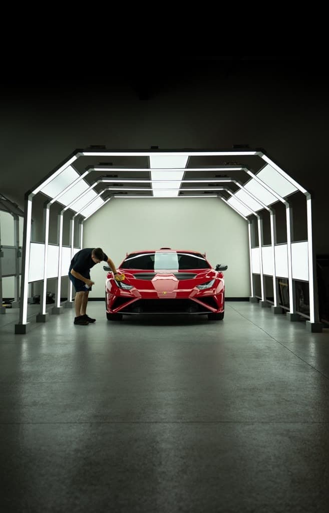 A Ferrari centered in the detail bay under full LED lighting, a detailer working the front bumper