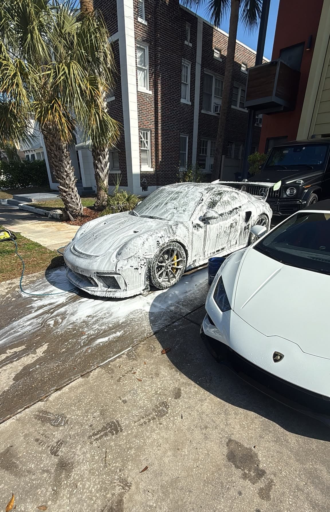 A coated sports car blanketed in snow foam during a maintenance wash, exotics parked alongside